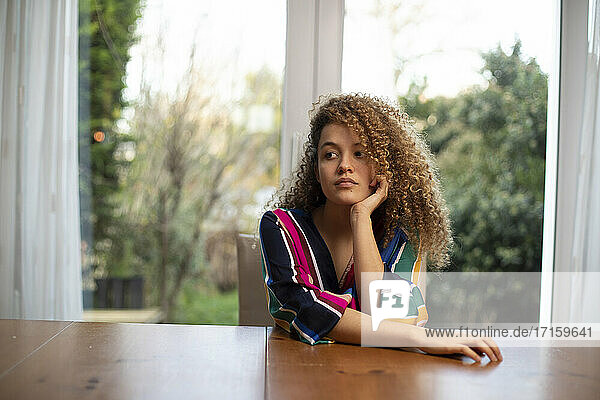 Nachdenkliche junge Frau sitzt am Tisch vor dem Fenster im Wohnzimmer
