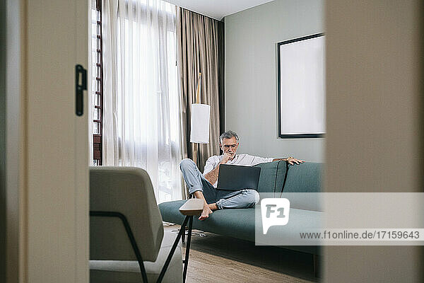 Thoughtful man looking at laptop while sitting on sofa in hotel room