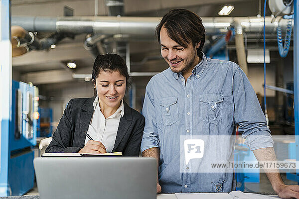 Smiling businesswoman working over laptop with colleague while standing at industry