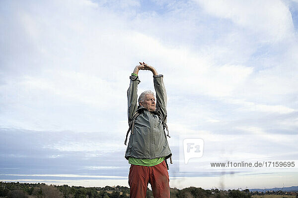 Senior male hiker stretching arms while standing against cloudy sky at countryside