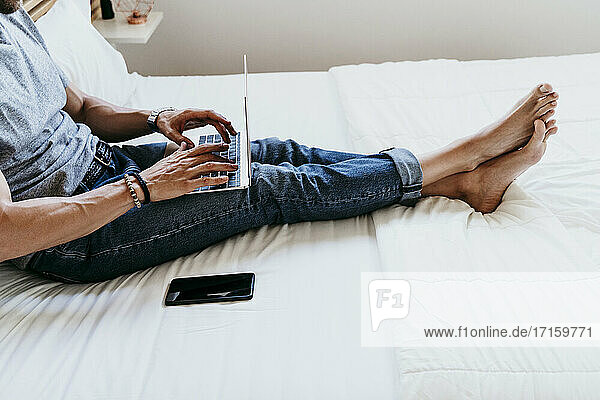 Junger Mann arbeitet mit Laptop und Mobiltelefon  während er zu Hause auf dem Bett sitzt
