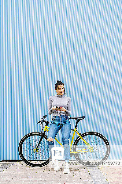 Woman using mobile phone looking away while sitting on fixie bike against blue wall