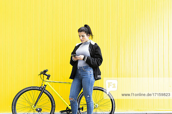 Schöne Frau  die ein Smartphone benutzt  während sie mit einem Fixie-Fahrrad vor einer gelben Wand steht