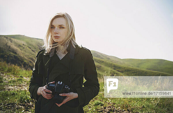 Beautiful blond woman with digital camera standing on hill against sky