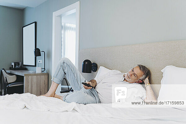 Relaxed man using remote control while lying on bed in hotel room