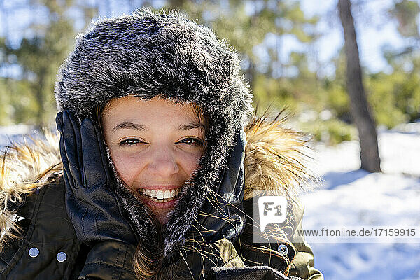 Cheerful woman wearing fur cap smiling while standing on forest during winter
