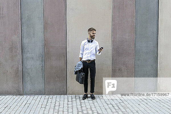 Businessman using smart phone while standing against wall
