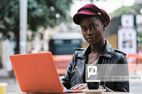 Young woman wearing jacket and cap using laptop while sitting at sidewalk cafe in city