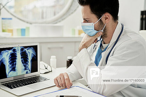 Thoughtful male doctor analyzing X-ray image on laptop while sitting in clinic