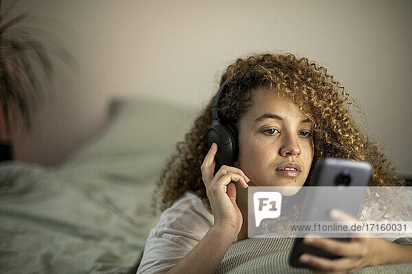 Young woman lying on bed while using smart phone and listening to music at home