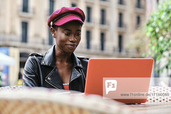 Smiling woman wearing cap working on laptop while sitting at sidewalk cafe