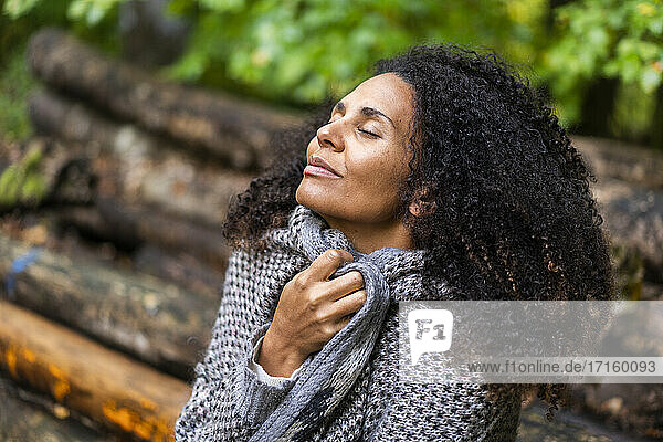 Frau mit lockigem Haar lächelt  während sie mit geschlossenen Augen im Wald sitzt
