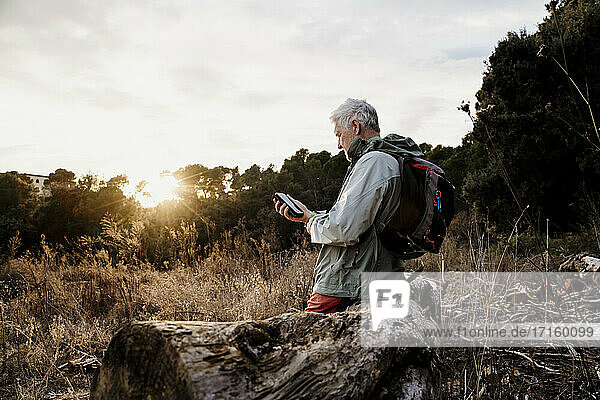 Älterer männlicher Wanderer  der am Wochenende auf einem Holzstamm sitzend ein Mobiltelefon benutzt