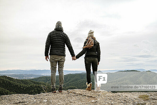 Couple holding hands while standing at viewpoint looking at mountains against sky