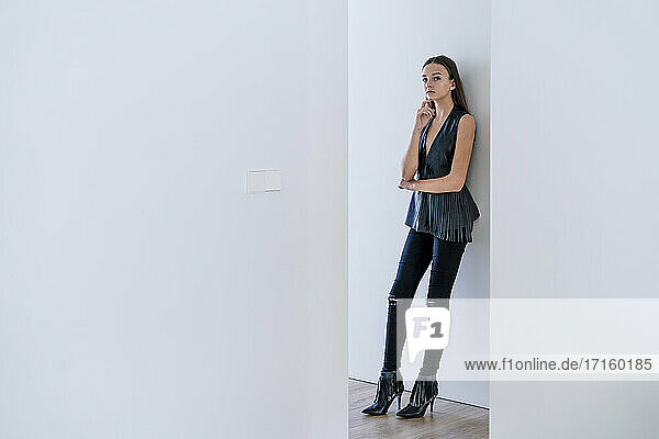 Teenage girl wearing stylish clothing leaning on wall at home seen through hallway