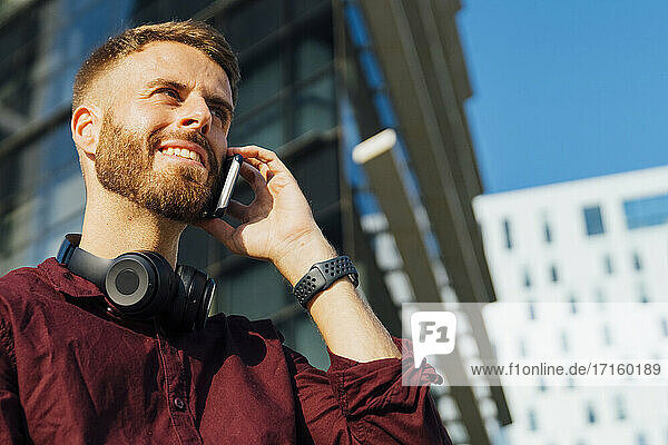 Lächelnder Geschäftsmann mit Kopfhörern  der in der Stadt mit seinem Mobiltelefon spricht