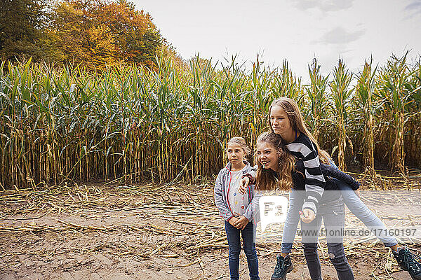 Smiling girl piggybacking friends while standing at corn field