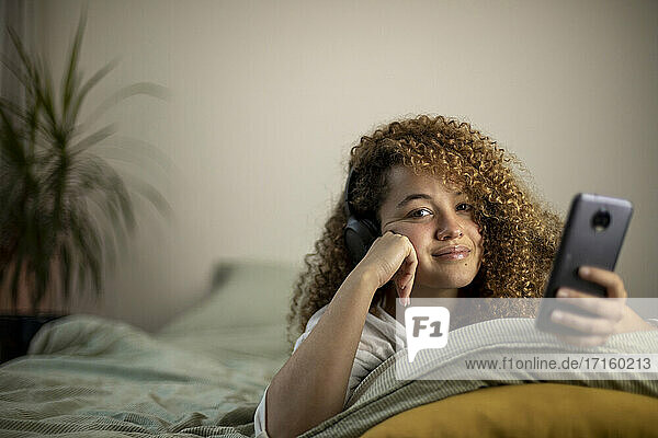 Smiling young woman with smart phone lying on bed in bedroom
