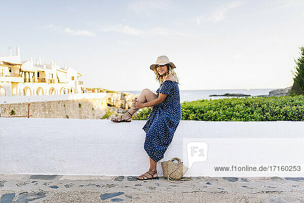 Weibliche Touristin mit Hut sitzt auf einer Stützmauer gegen den Himmel im Dorf Binibeca  Menorca  Spanien