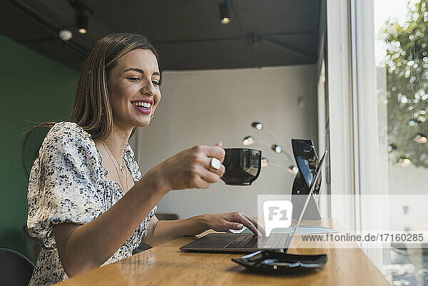 Glückliche Unternehmerin mit Kaffee in der Hand und Laptop auf dem Tisch im Café