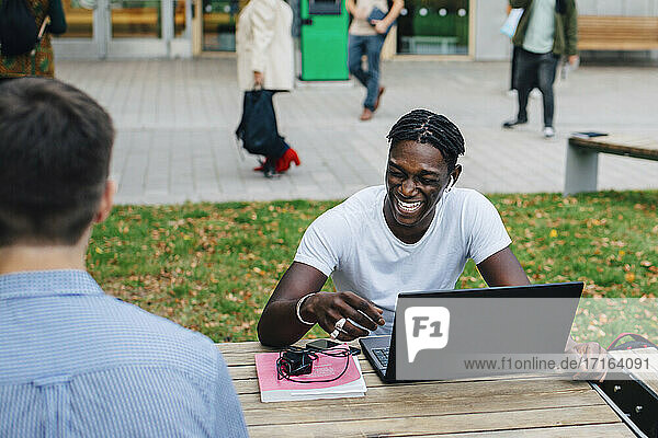 Junger männlicher Student lachend beim Sitzen mit Freund in Campus