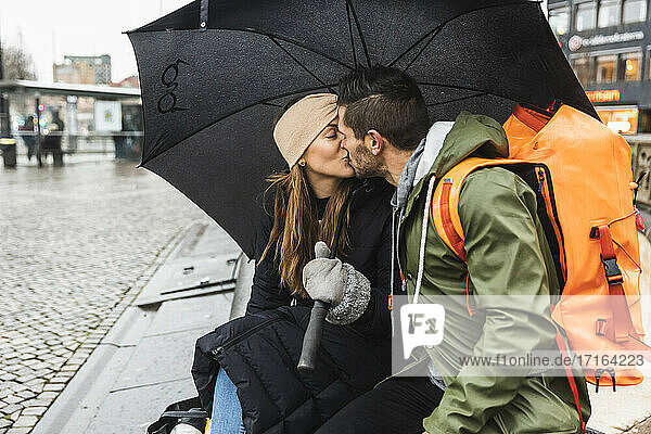 Männlich und weiblich Tourist küssen einander beim Sitzen auf dem Bürgersteig während der Regenzeit