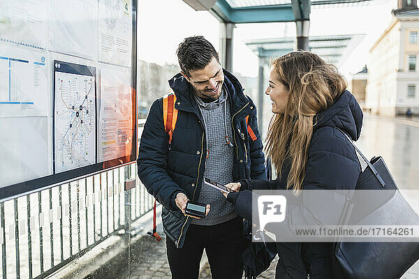 Lächelnde Frau scannt QR-Code durch Smartphone steht neben Mann am Bahnhof