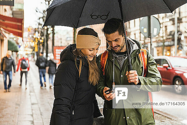 Lächelndes Paar mit Smartphone beim Gehen auf dem Fußweg in der Stadt während der Regenzeit