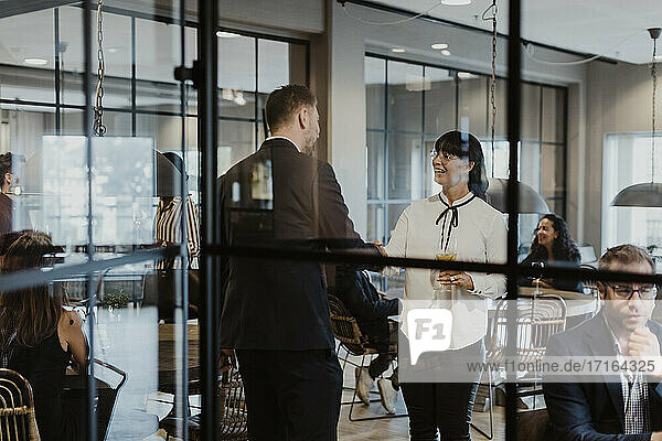 Geschäftsfrau schüttelt Hände mit männlichen Kollegen durch Glas während gesellschaftlicher Veranstaltung im Büro gesehen