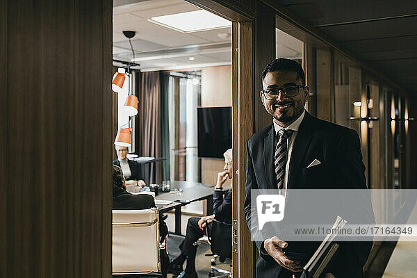 Porträt eines lächelnden männlichen Berufstätigen  der Dokumente hält  während er sich an die Tür zum Vorstandszimmer im Büro lehnt