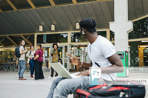 Männlicher Student  der seine Freunde anschaut  während er einen Laptop auf dem Campus benutzt