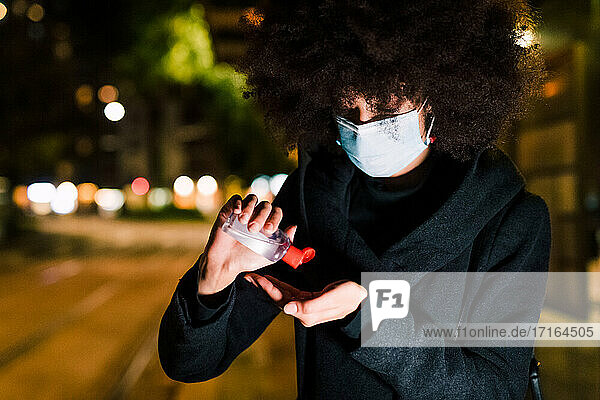 Junge Frau trägt eine Gesichtsmaske und trägt Handdesinfektionsmittel auf  nachts im Freien