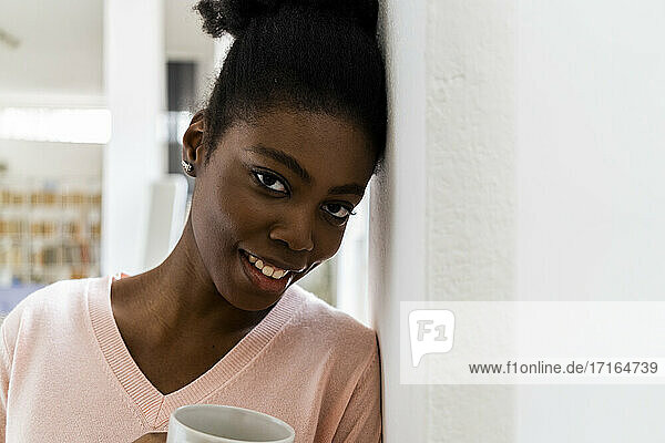Smiling woman with coffee cup staring while leaning in wall at home
