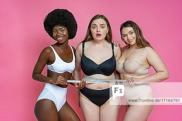Lächelnde multiethnische weibliche Modelle in Dessous  die die Taille einer besorgten übergroßen Freundin messen  vor einem rosa Hintergrund