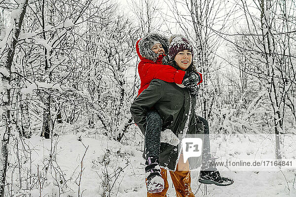 Teenager-Mädchen gibt Huckepack zu Schwester gegen Schnee bedeckt kahlen Bäumen im Wald