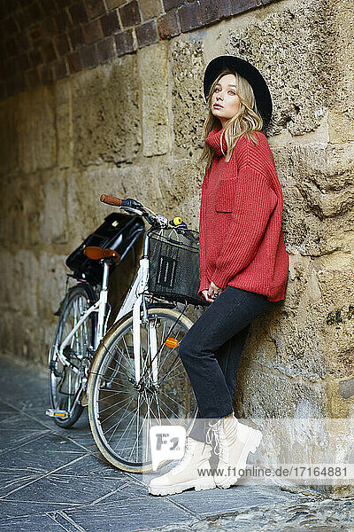 Young woman wearing hat looking away while leaning on wall by bicycle