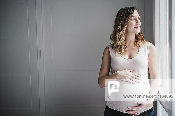 Nachdenkliche schwangere Frau mit Händen auf dem Bauch  die durch ein Fenster zu Hause schaut