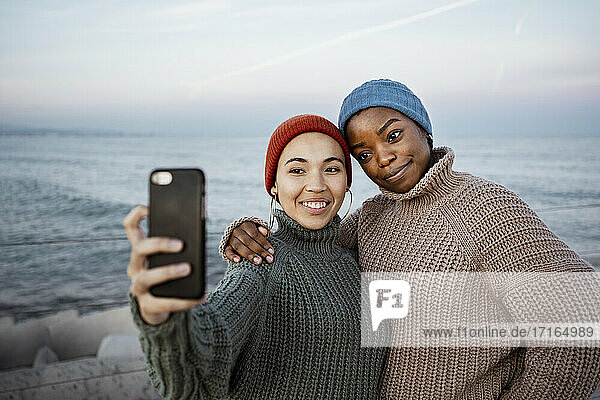 Freunde mit Strickmütze  die lächelnd ein Selfie mit dem Handy machen  während sie am Meer stehen