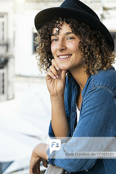Smiling young woman with hat leaning on handrail of balcony