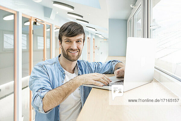 Glückliche kreative männliche Fachkraft mit Laptop im Flur am Arbeitsplatz stehend