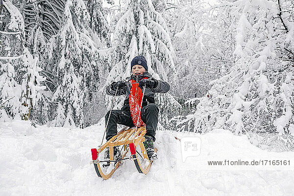 Playful boy sledding while sitting on sled in forest during winter