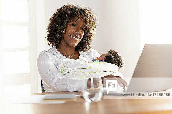 Lächelnde Mutter mit Baby sitzend bei der Arbeit am Laptop zu Hause