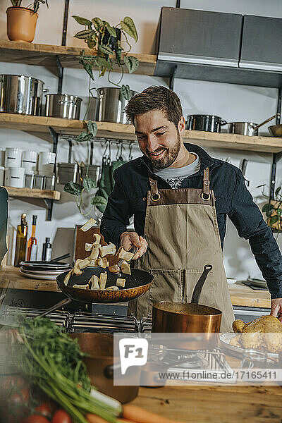 Smiling male chef tossing mushroom in frying pan while standing in kitchen