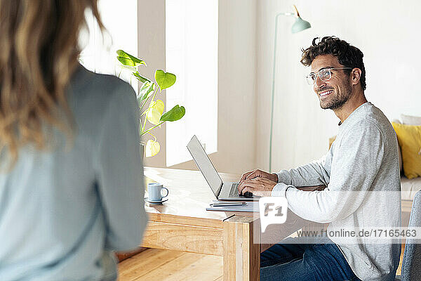 Lächelnder Mann  der eine Frau ansieht  während er im Büro zu Hause am Laptop arbeitet