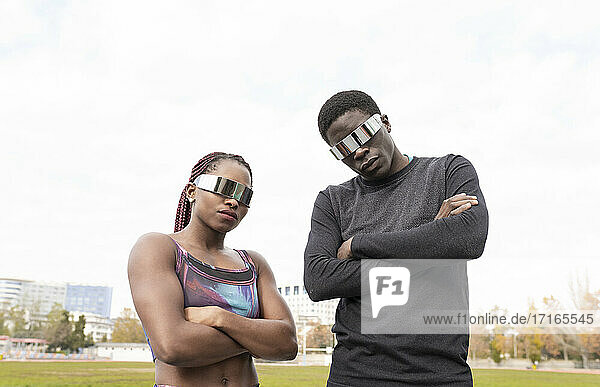 Junge Sportlerin und junger Sportler mit Sonnenbrille und verschränkten Armen vor einem klaren Himmel