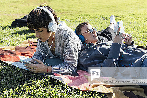 Young friends wearing headphones using smart phones while lying on blanket at park