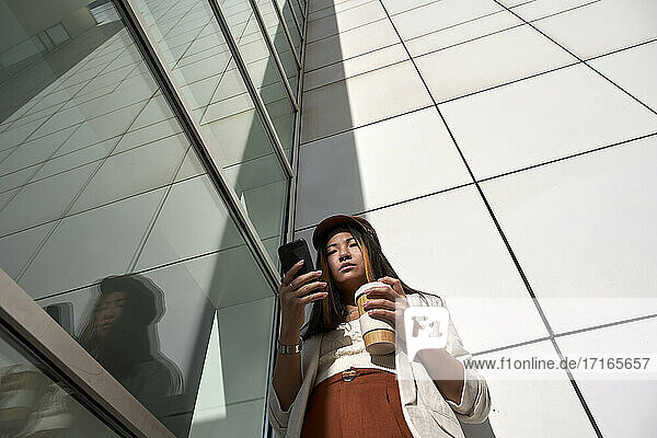 Beautiful woman with reusable cup using smart phone while standing against wall