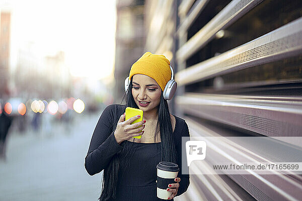Beautiful woman with reusable cup using smart phone by railing at street