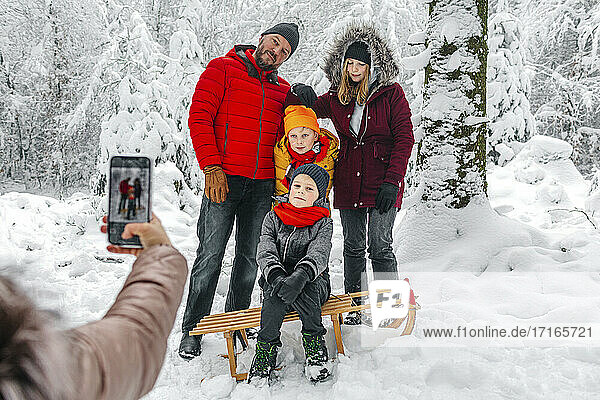 Woman taking photo of family through mobile phone while standing in forest during winter