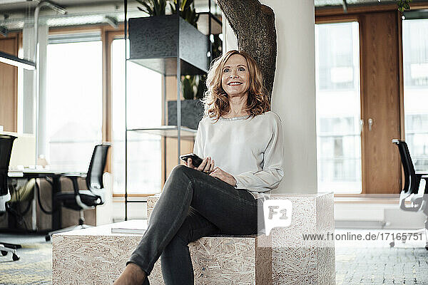 Smiling businesswoman with smart phone sitting on retaining wall in creative office